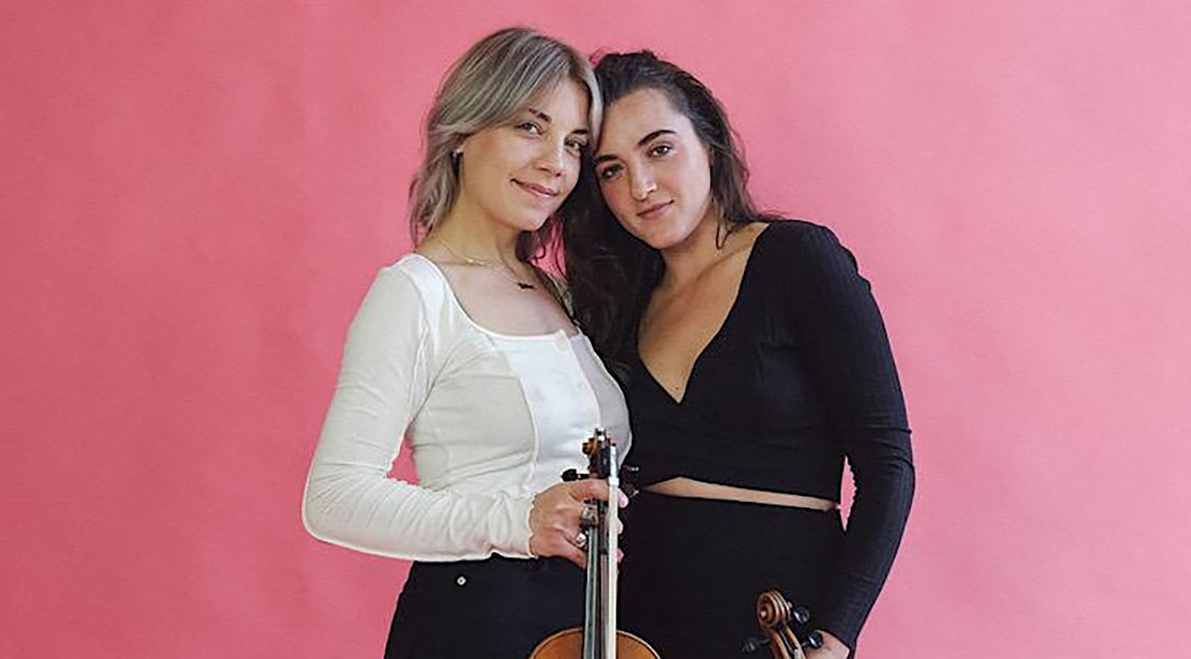 two women holding string instruments in front of a pink wall